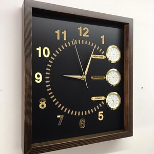 May include: A square wooden framed clock with a black face and gold numbers. The clock has three smaller clocks on the face, each with a different city name: New York, Buffalo, and Sydney.