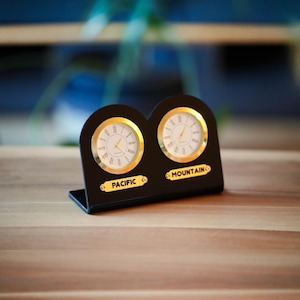 May include: A black dual-dial clock with gold-colored accents. Each clock face has a white background with black Roman numerals and gold hands. The base has the words "PACIFIC" and "MOUNTAIN" in gold lettering.