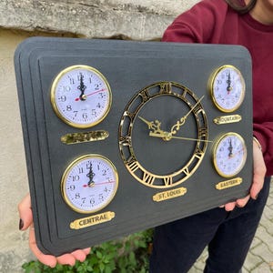 Könnte beinhalten: Eine dunkelgraue Mehrzeitzonenuhr mit fünf Zifferblättern. Die zentrale Uhr hat goldene römische Ziffern und Zeiger. Die kleineren Uhren sind mit "Central", "Mountain" und "Eastern" beschriftet.