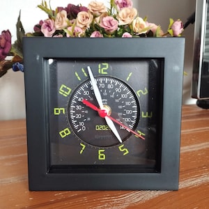 Könnte beinhalten: Eine schwarze quadratische Uhr mit weißem Zifferblatt und schwarzen Zeigern. Das Zifferblatt der Uhr hat ein Tachodesign mit Zahlen in Grün und Rot. Das Zifferblatt der Uhr enthält auch den Text "Kirpiirt" und "MPH".
