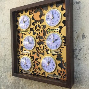 May include: A wooden framed wall clock with gold gears and six smaller clocks displaying different time zones. The time zones are labeled: Pacific, Mountain, Eastern, Central, Hawaii, and London.