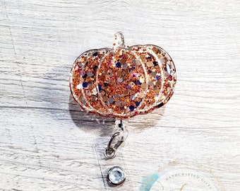 Può includere: Un portabadge retrattile a forma di zucca con glitter dorati, un portabadge in plastica trasparente e una clip in metallo argentato. Il portabadge è decorato con glitter dorati, arancioni e marroni. Il testo "Sidney Lynn Designs Made Especially For You" è visibile sul portabadge.