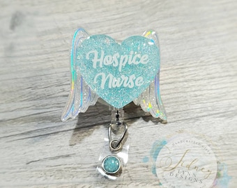 May include: A badge reel with a heart-shaped design and angel wings. The heart is light blue with glitter and the words "Hospice Nurse" in white script. The wings have an iridescent sheen.