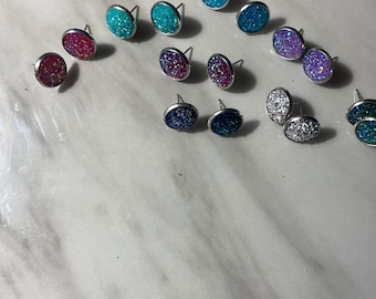 May include: A collection of oval-shaped stud earrings in various colors, including teal, purple, silver, and dark blue. Each earring has a glittery, textured surface and a silver post. The earrings are arranged on a white surface.
