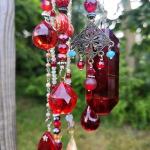 May include: A decorative suncatcher featuring multiple strands of red glass beads, faceted red crystal pendants, and a central antique bronze-toned filigree charm. The assembly includes small turquoise accents, a metallic cone, and clear beads.