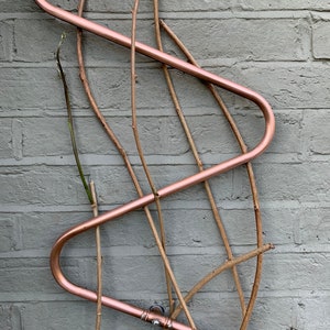 May include: A copper trellis with a wavy design is attached to a brick wall.  The trellis is designed to support climbing plants.  The trellis is made of copper tubing and is attached to the wall with screws.