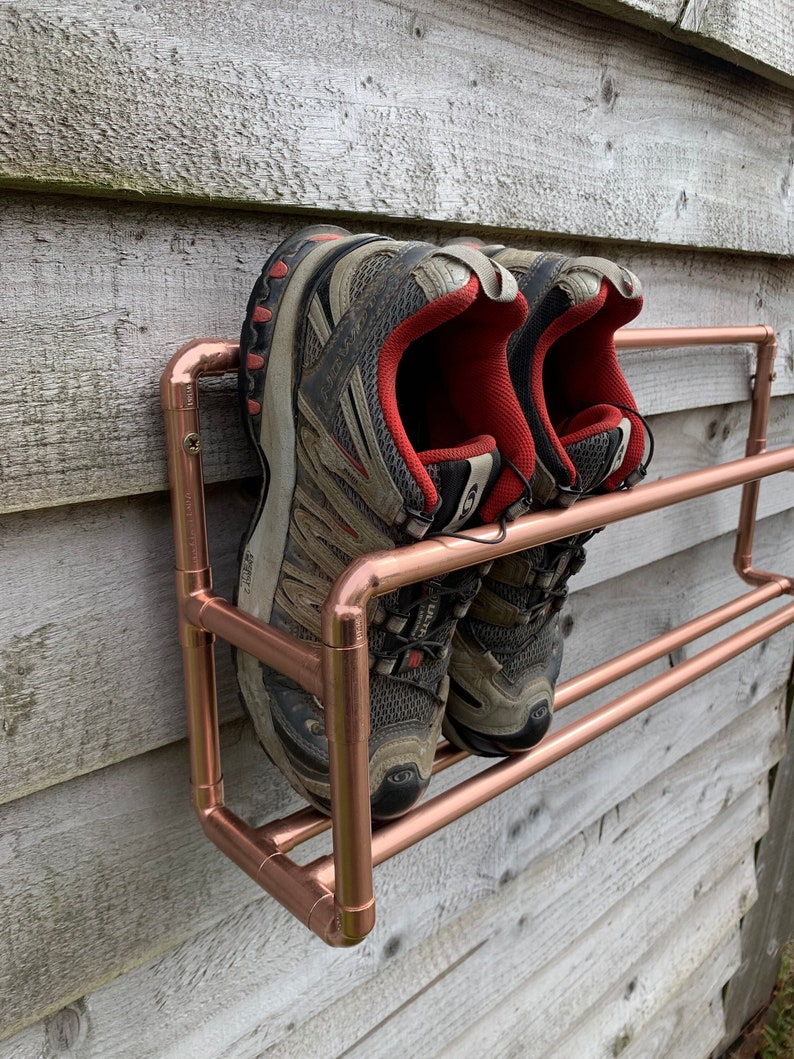 May include: A copper pipe shoe rack mounted on a wooden wall with two pairs of black and red sneakers on the rack.