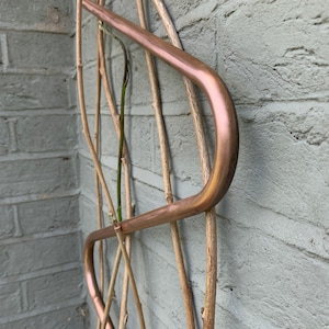 May include: A copper plant trellis with a curved design. The trellis is attached to a brick wall and has several wooden branches woven through it.