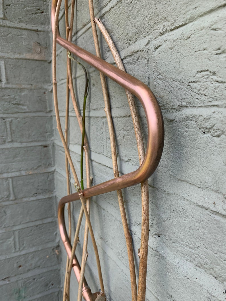May include: A copper plant trellis with a curved design. The trellis is attached to a brick wall and has several wooden branches woven through it.