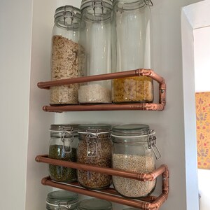May include: Copper pipe shelving unit with glass jars filled with various dry goods, such as pasta, rice, lentils, and beans. The shelves are mounted on a white wall.