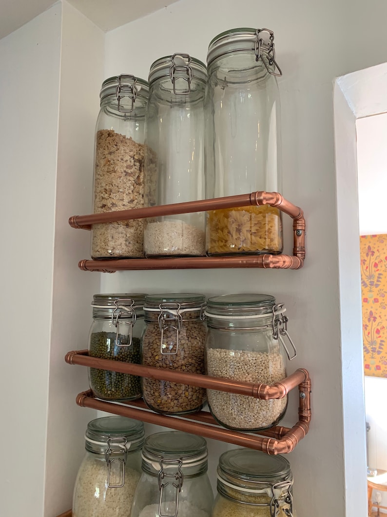 May include: Copper pipe shelving unit with glass jars filled with various dry goods, such as pasta, rice, lentils, and beans. The shelves are mounted on a white wall.