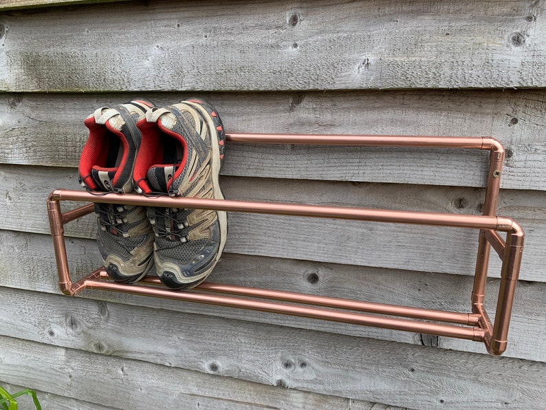 May include: A copper pipe shoe rack with two shelves mounted on a wooden wall. Two pairs of black and gray hiking shoes are on the top shelf.