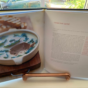 May include: A close-up of an open cookbook with a recipe for chicken liver parfait. The recipe includes a list of ingredients and instructions. The image also shows a bowl of chicken liver parfait with a garnish of green leaves.