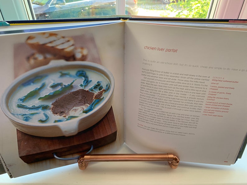 May include: A close-up of an open cookbook with a recipe for chicken liver parfait. The recipe includes a list of ingredients and instructions. The image also shows a bowl of chicken liver parfait with a garnish of green leaves.