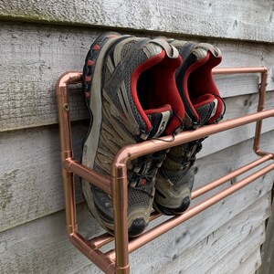 May include: A copper pipe shoe rack with two shelves mounted on a wooden wall. Two pairs of gray and red athletic shoes are on the shelves.