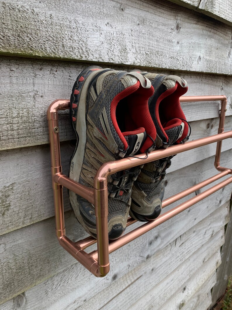 May include: A copper pipe shoe rack with two shelves mounted on a wooden wall. Two pairs of gray and red athletic shoes are on the shelves.