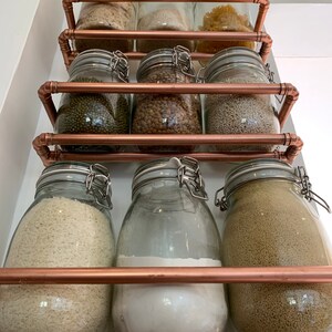 May include: A copper pipe shelving unit with glass jars filled with various dry goods, such as rice, lentils, and pasta. The jars have metal lids and are arranged in three rows.