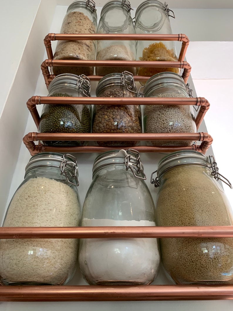 May include: A copper pipe shelving unit with glass jars filled with various dry goods, such as rice, lentils, and pasta. The jars have metal lids and are arranged in three rows.
