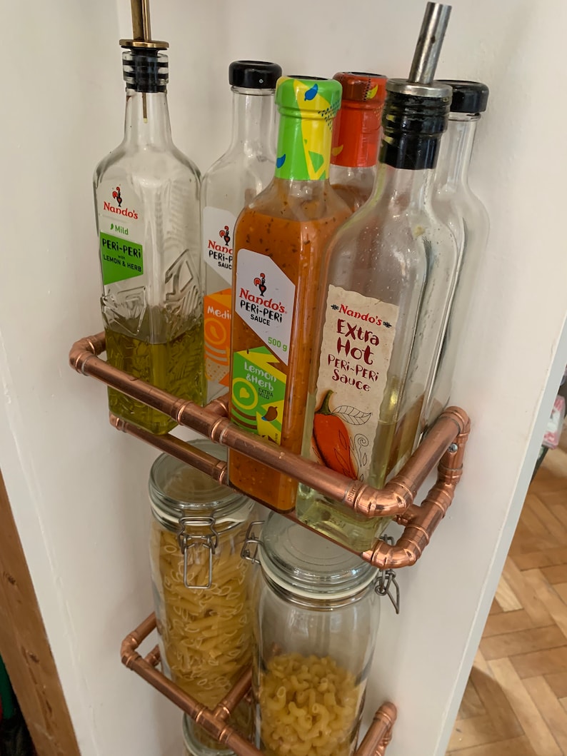 May include: A copper pipe shelf with four glass jars filled with pasta and five glass bottles with Nando's Peri-Peri sauce. The bottles are labeled with different flavors, including Mild, Lemon & Herb, and Extra Hot.