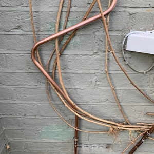 May include: A copper pipe bent into a curved shape is attached to a brick wall. The pipe is surrounded by several thin, dried branches.