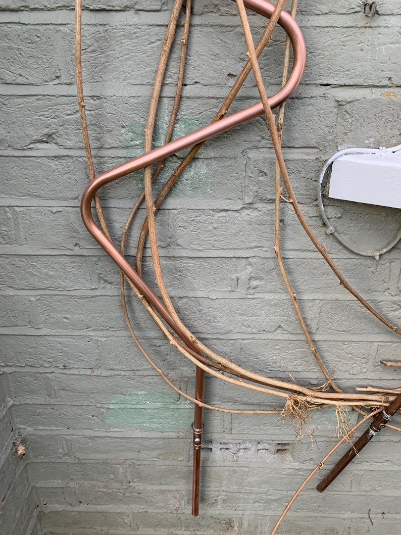 May include: A copper pipe bent into a curved shape is attached to a brick wall. The pipe is surrounded by several thin, dried branches.