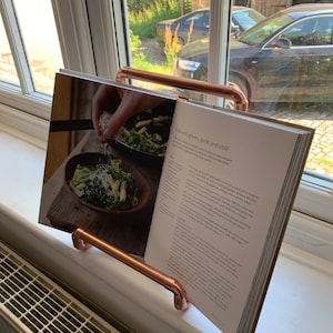 May include: A copper pipe book stand holding a cookbook open to a recipe for pasta with greens, garlic and chili. The recipe includes instructions for cooking the pasta and adding the greens, garlic, chili and parmesan cheese.