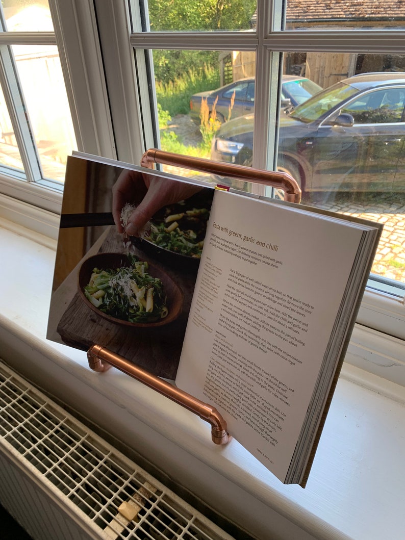 May include: A copper pipe book stand holding a cookbook open to a recipe for pasta with greens, garlic and chili. The recipe includes instructions for cooking the pasta and adding the greens, garlic, chili and parmesan cheese.