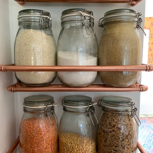 May include: A set of six glass jars with airtight lids, filled with various dry goods, are organized on a copper pipe shelf. The jars are filled with rice, flour, couscous, lentils, split peas, and brown sugar.