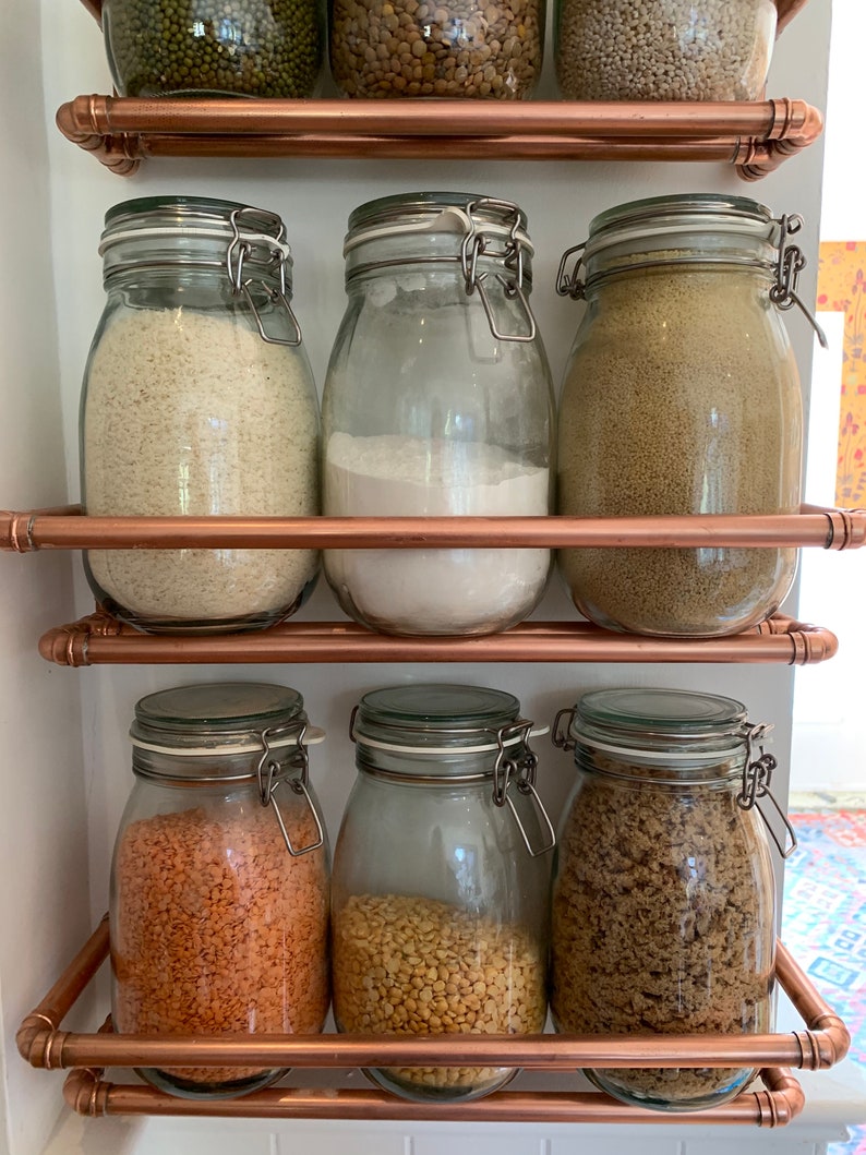 May include: A set of six glass jars with airtight lids, filled with various dry goods, are organized on a copper pipe shelf. The jars are filled with rice, flour, couscous, lentils, split peas, and brown sugar.