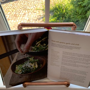 May include: Open cookbook with a recipe for pasta with greens, garlic and chili. The recipe includes a photo of a bowl of pasta with greens and grated cheese. The cookbook is propped open on a copper pipe stand.