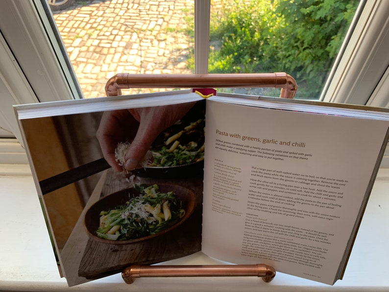 May include: Open cookbook with a recipe for pasta with greens, garlic and chili. The recipe includes a photo of a bowl of pasta with greens and grated cheese. The cookbook is propped open on a copper pipe stand.