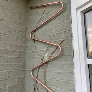 May include: A copper pipe trellis attached to a brick wall. The trellis is shaped like a zig-zag and is designed to support climbing plants.