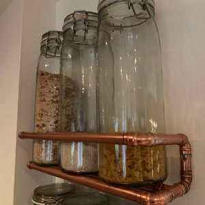 May include: Four glass jars with metal lids are filled with various dry goods and are displayed on a copper pipe shelf. The shelf is mounted on a white wall.