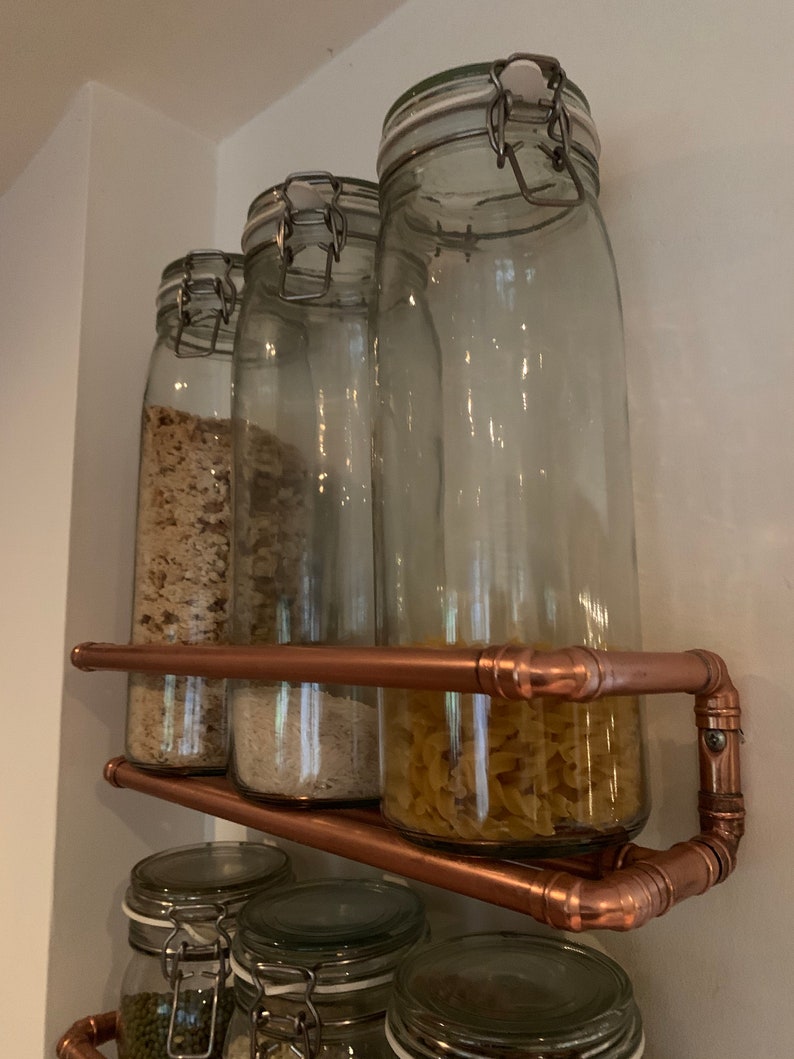 May include: Four glass jars with metal lids are filled with various dry goods and are displayed on a copper pipe shelf. The shelf is mounted on a white wall.