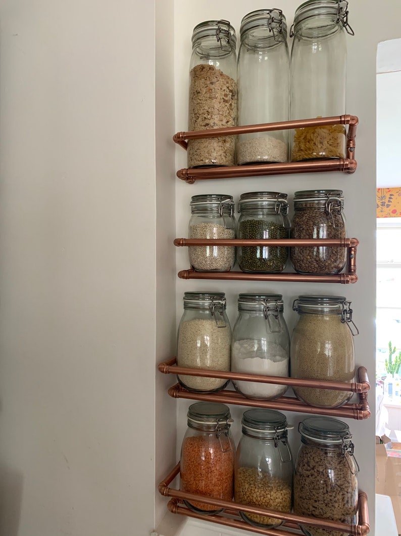 May include: A copper pipe shelving unit with glass jars filled with various dry goods, such as grains, beans, and pasta. The jars have metal lids with clips.