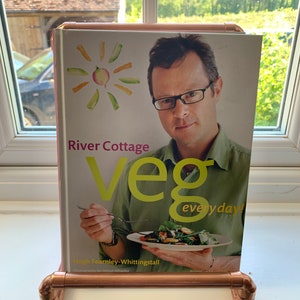 May include: A pink and white cookbook titled "River Cottage Veg everyday!" by Hugh Fearnley-Whittingstall. The book is displayed on a copper pipe book stand.