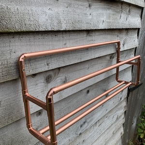 May include: A copper towel rack mounted on a wooden wall. The rack has two bars and is made of copper pipe.