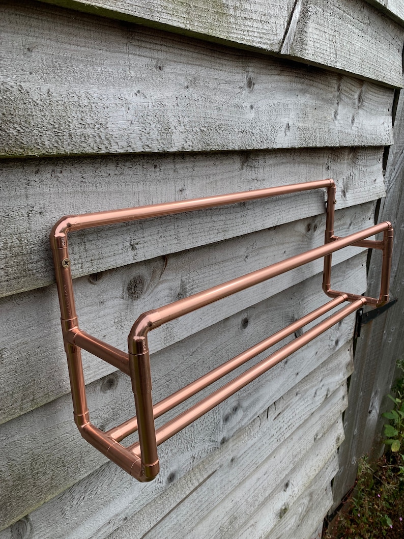 May include: A copper towel rack mounted on a wooden wall. The rack has two bars and is made of copper pipe.