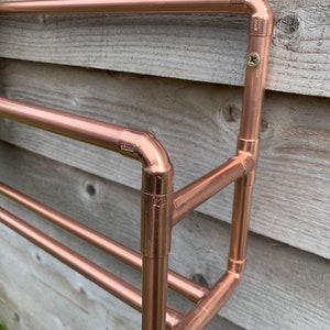 May include: A copper pipe shoe rack mounted on a wooden wall. The rack has three shelves and is made of copper pipes and fittings.
