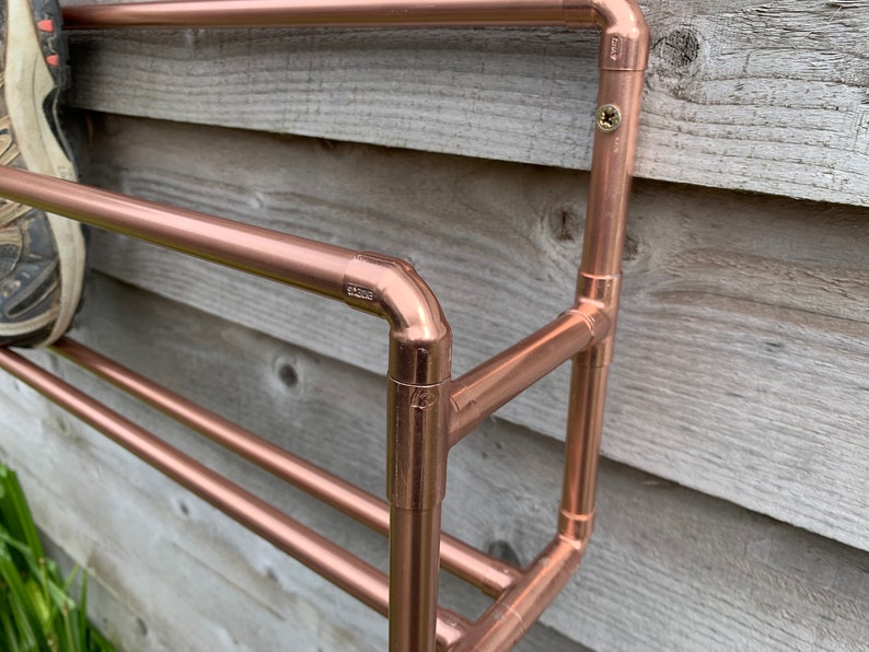 May include: A copper pipe shoe rack mounted on a wooden wall. The rack has three shelves and is made of copper pipes and fittings.