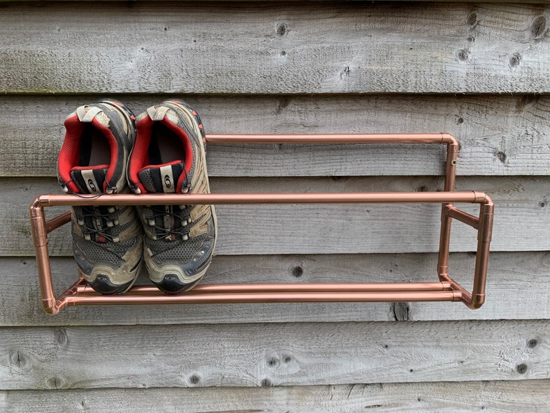 May include: A two-tier copper pipe shoe rack mounted on a wooden wall. Two pairs of gray and black hiking shoes are on the rack.