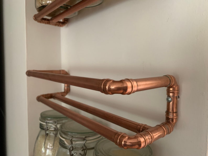 May include: Copper pipe shelving unit with two shelves. The shelves are made of copper pipe and are attached to the wall with copper pipe brackets. The shelves are empty.