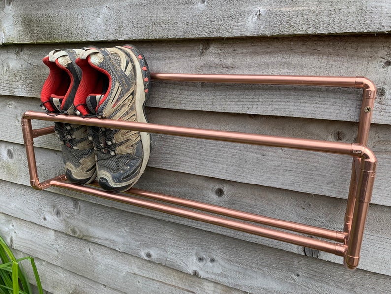 May include: A copper pipe shoe rack with two shelves mounted on a wooden wall. Two pairs of black and red sneakers are on the top shelf.