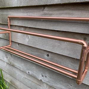 May include: A copper pipe shelf with two tiers mounted on a wooden wall. The shelf is made of copper pipe and fittings.