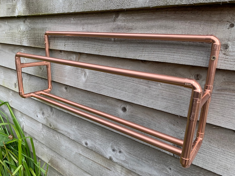May include: A copper pipe shelf with two tiers mounted on a wooden wall. The shelf is made of copper pipe and fittings.