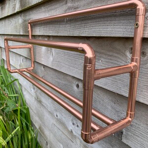 May include: A copper pipe shelf with two tiers, mounted on a wooden wall. The shelf is made of copper pipe and fittings, and has a polished finish.
