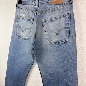 May include: Light blue denim jeans with a classic five-pocket design, visible back pockets, and a leather patch. The jeans are hanging on a black hanger, showcasing the back view of the garment. The denim has a faded, worn look.
