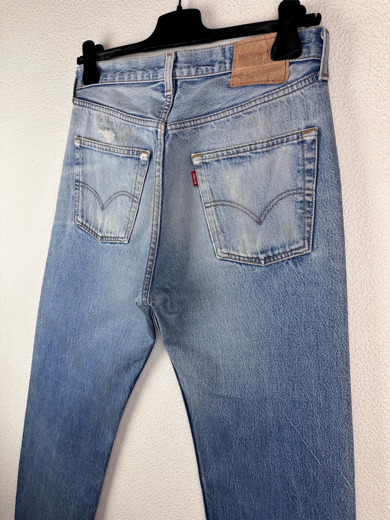 May include: Light blue denim jeans with a classic five-pocket design, visible back pockets, and a leather patch. The jeans are hanging on a black hanger, showcasing the back view of the garment. The denim has a faded, worn look.