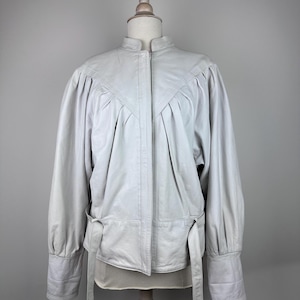 Peut inclure: Veste en cuir gris clair avec col montant et manches longues. La veste présente une taille cintrée avec une ceinture et des détails plissés aux épaules. Elle est présentée sur un mannequin en bois. Mesures en centimètres.