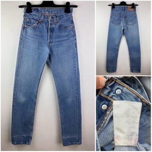 Könnte beinhalten: Hellblaue Jeans mit Knopfleiste, fünf Taschen und verwaschener Optik. Die Jeans werden auf einem Kleiderbügel präsentiert, der die Vorder- und Rückseite zeigt. Eine Nahaufnahme zeigt das Etikett und die Details des Denims.